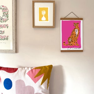 May include: A framed print of a cheetah with a pink background and the text "El Leopardo". The print is hanging on a wall with a white pillow with a geometric pattern in front of it.