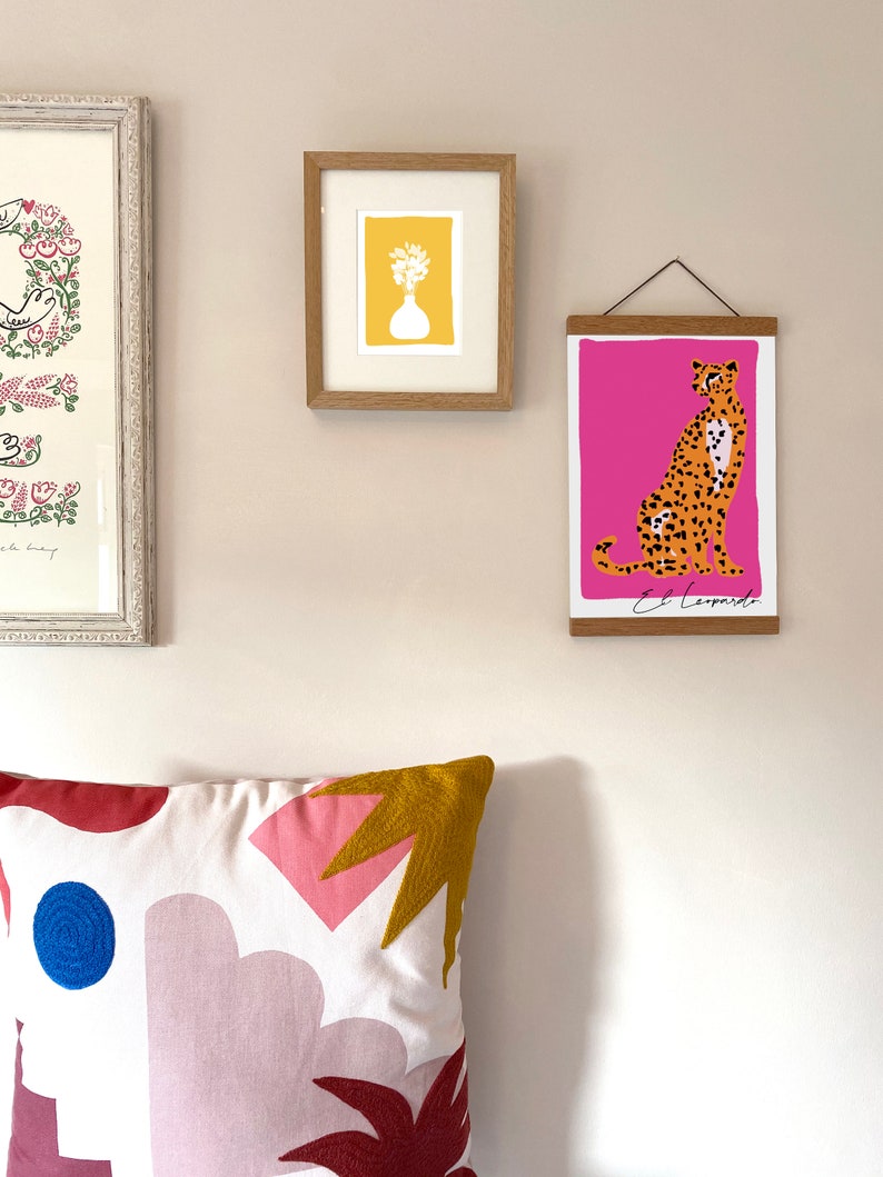 May include: A framed print of a cheetah with a pink background and the text "El Leopardo". The print is hanging on a wall with a white pillow with a geometric pattern in front of it.