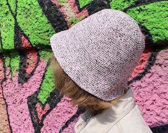 May include: A light pink and black knit bucket hat. The hat is worn by a person in a tan jacket. The background is a wall with graffiti art in green, pink, and black.