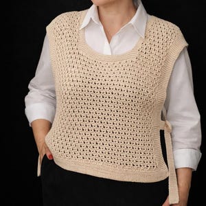 May include: A beige sleeveless crochet vest featuring an openwork stitch pattern, a wide scoop neckline, and solid knit borders. The vest has side openings secured with matching knit ties at the waist, creating a layered look over a white shirt.