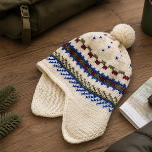 May include: A cream-colored knit hat with ear flaps and a pom-pom on top. The hat features a horizontal pattern of blue, green, and maroon geometric shapes and zig-zag lines encircling the crown, crafted from thick yarn with a textured finish.