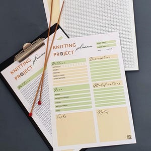 May include: A knitting project planner printed on white paper with green and yellow sections, featuring fields for pattern, yarn, tasks, and notes. Two brown knitting needles with red tips are placed diagonally across the planner pages.
