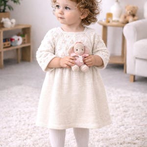 May include: A young child wearing a cream-colored knitted dress, white tights, and burgundy Mary Jane shoes. The child is holding a small pink stuffed bunny. The background includes a white rug and wooden furniture.