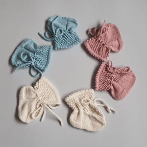 May include: Six hand-knitted baby booties arranged in a circle. The set includes two light blue booties, two dusty rose pink booties, and two cream-coloured booties. Each pair features a textured knit pattern on the cuff and a matching tie closure.