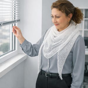 May include: A white triangular shawl featuring a delicate, open-knit lace pattern. The shawl is draped around the neck and shoulders, with its pointed ends hanging down the front. The fabric has a sheer, lightweight appearance with visible texture.