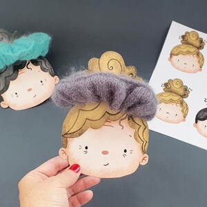 May include: Two illustrated paper cutouts of faces with fuzzy hair accessories, one in teal and one in dark grey, featuring gold-toned hair details. A white sheet with four additional printed face illustrations is visible in the background.