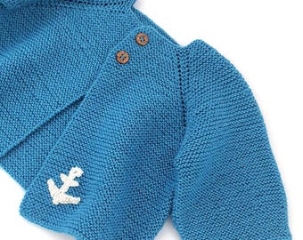 May include: A bright blue knitted cardigan featuring long sleeves, a folded collar, and two brown wooden buttons at the neckline. A small, white, crocheted anchor applique is attached to the lower front corner of the garment.