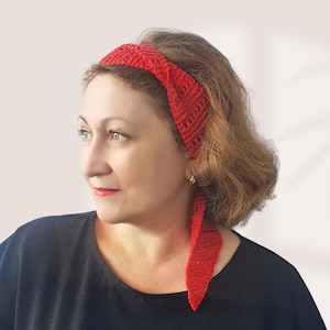 May include: A bright red crochet headband featuring a textured, open-knit pattern with a twisted knot detail at the top and a long, pointed tail extending down one side, crafted from yarn with a visible stitch structure throughout the accessory.
