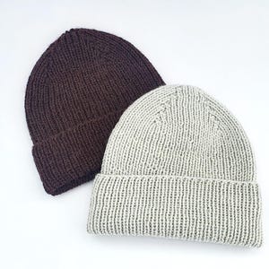 Puede incluir: Dos gorros de punto, uno de color marrón oscuro y otro de color gris claro. Ambos gorros presentan una textura acanalada, un dobladillo plegado en la parte inferior y una corona redondeada. Los gorros están colocados uno al lado del otro.