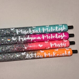 May include: Four glitter pens in teal, pink, coral, and orange. Each pen has a name written in white script with small white hearts. The pens have black tips and are arranged horizontally.