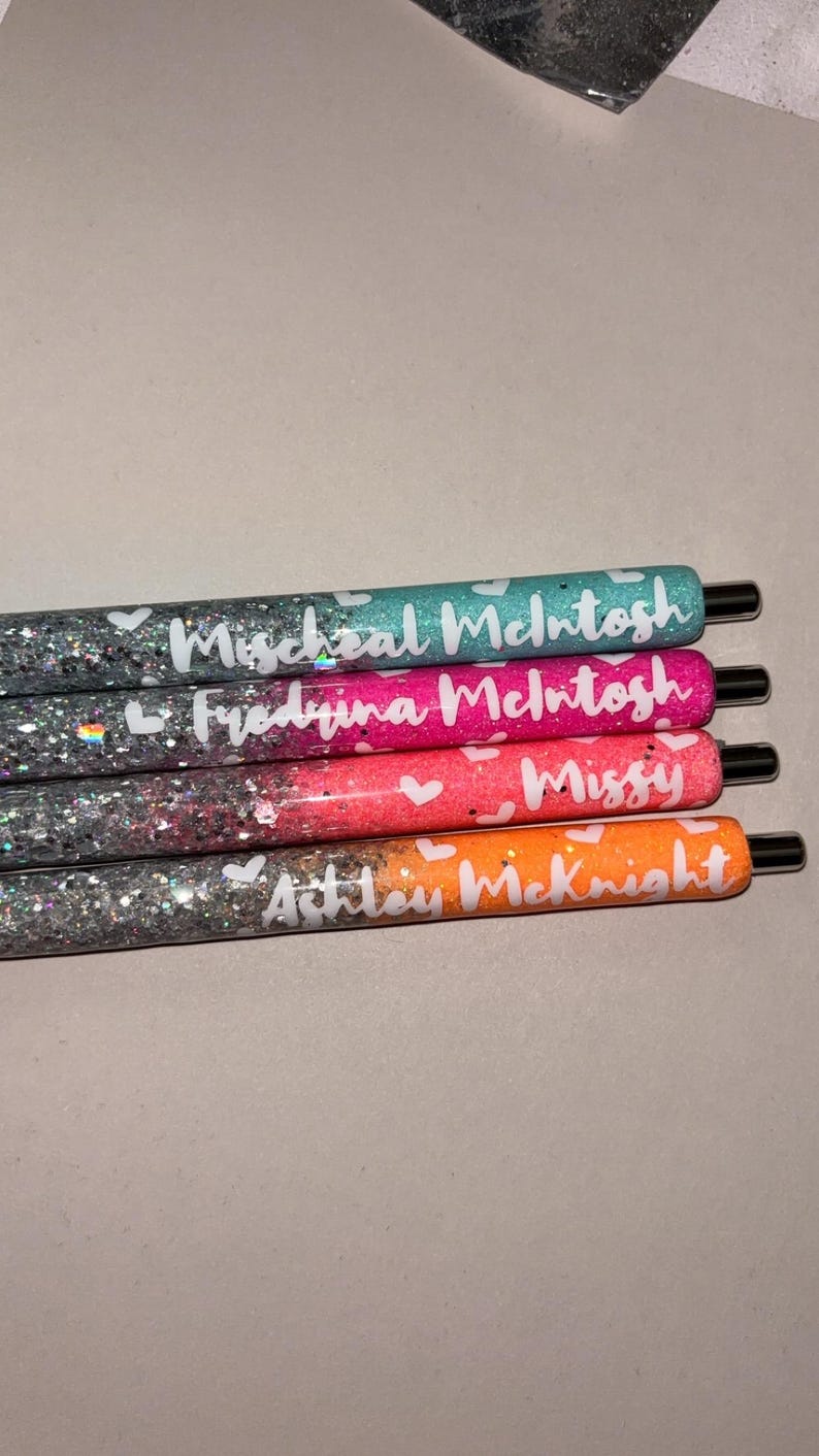 May include: Four glitter pens in teal, pink, coral, and orange. Each pen has a name written in white script with small white hearts. The pens have black tips and are arranged horizontally.