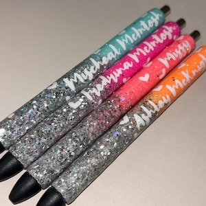 May include: Four glitter pens in teal, pink, orange, and silver. Each pen has a different name in white script: "Micheal McIntosh", "Medryna McIntosh", "Missy", and "Ashley McKnight". The pens have black tips.
