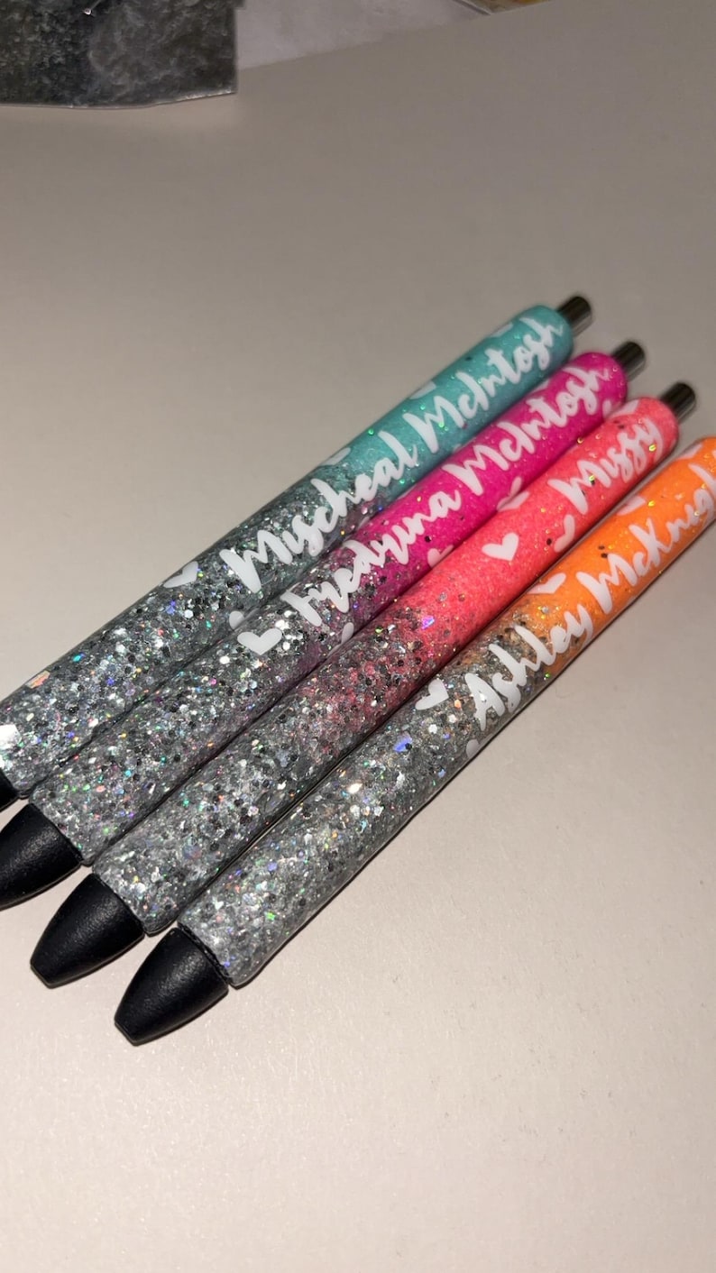 May include: Four glitter pens in teal, pink, orange, and silver. Each pen has a different name in white script: "Micheal McIntosh", "Medryna McIntosh", "Missy", and "Ashley McKnight". The pens have black tips.