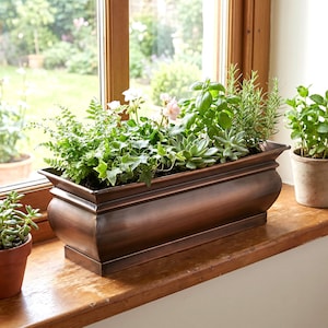 May include: A rectangular, bronze-colored planter box filled with various green plants and small flowers. The planter sits on a wooden windowsill in front of a window with a view of a garden. The planter has a decorative, curved design.