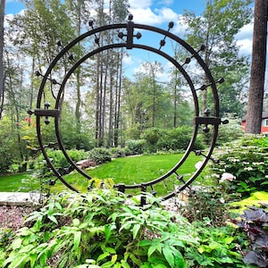 May include: A large, dark metal garden arch with decorative finials stands in a lush green garden. The arch is circular, with a detailed design. The background features a vibrant lawn, trees, and a glimpse of a red house under a blue sky.