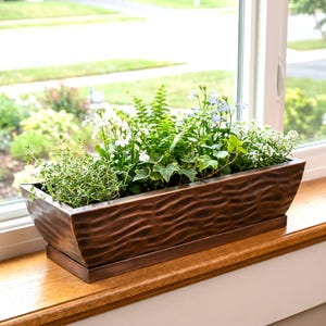 May include: A rectangular, copper-colored metal planter box filled with various green plants and white and blue flowers. The planter sits on a wooden windowsill in front of a window with a view of a green lawn and trees.