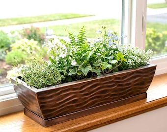 May include: A rectangular, copper-colored metal planter box filled with various green plants and white and blue flowers. The planter sits on a wooden windowsill in front of a window with a view of a green lawn and trees.