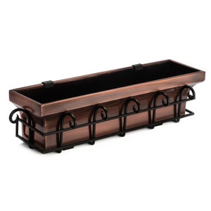 May include: A copper-colored window box planter with a black metal frame. The frame has decorative scrollwork and brackets for mounting.