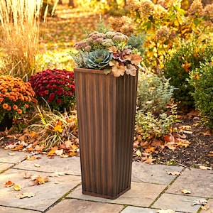 May include: A tall, square, bronze-colored planter with vertical fluted sides. The planter is filled with a variety of plants, including a central green cabbage-like succulent, pinkish-brown flowering clusters, and various green foliage.
