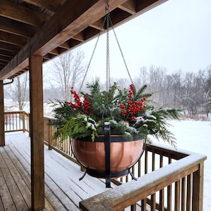 May include: A copper hanging planter bowl held by a black metal frame and chains, filled with green pine branches, cedar sprigs, and bright red berries. The arrangement is dusted with a light layer of snow, creating a seasonal display.