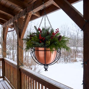 May include: A polished copper bowl planter suspended by a black metal frame and chains, filled with evergreen boughs, pinecones, and bright red berries, hanging from a wooden porch ceiling overlooking a snowy landscape.