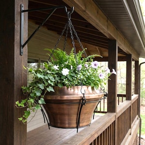May include: A copper-colored hanging planter filled with green foliage and purple and white flowers. The planter is suspended by a black chain from a black metal bracket attached to a wooden post. The scene is set on a porch.