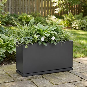May include: A rectangular, dark gray planter box filled with various green plants and white flowers. The planter sits on a matching tray, and is placed on a stone patio with a wooden fence in the background.