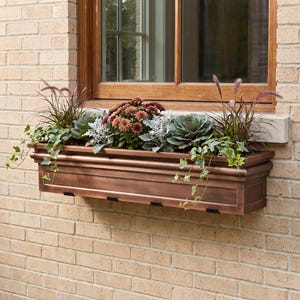 May include: A rectangular copper-toned window box planter mounted against a light-colored brick wall. The planter is filled with a variety of plants, including dark red chrysanthemums, green ornamental kale, trailing ivy, and tall purple grasses.