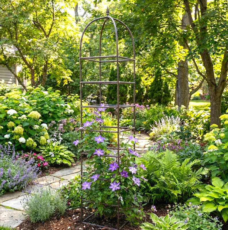 May include: A tall, dark metal garden obelisk trellis with a pointed finial at the top, featuring a square grid structure. The trellis is positioned in a garden, with purple clematis flowers climbing up the lower sections of the frame.