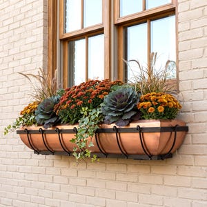 May include: A rectangular copper window box planter with a black metal frame, filled with orange chrysanthemums, purple ornamental kale, green ivy, and tall ornamental grasses, mounted on a light-colored brick wall beneath a wooden window.