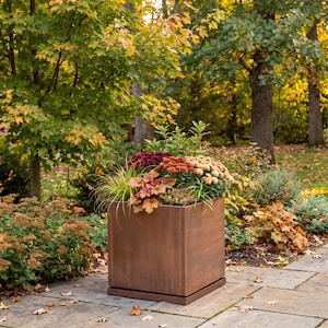 May include: A square, metallic planter with a brushed copper finish and a matching base tray. The planter is filled with a variety of autumn-colored flowers, including deep red, orange, and cream-colored chrysanthemums and green foliage.
