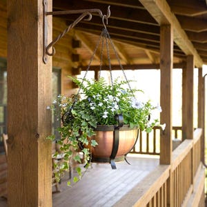 May include: A copper-colored hanging planter filled with green foliage and white flowers. The planter is suspended by chains from a decorative wrought iron hook attached to a wooden post. The porch setting has a rustic aesthetic.