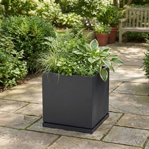 May include: A square, dark gray planter box filled with various green plants and foliage. The planter sits on a matching tray, and is placed on a stone patio. The background includes a garden with other plants and a wooden bench.