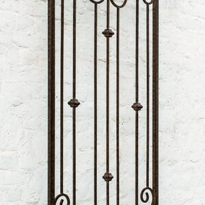 Pu&ograve; includere: Un alto traliccio da parete rettangolare in metallo marrone scuro con una parte superiore ad arco. La struttura &egrave; composta da barre verticali arricchite da piccole sfere decorative e complessi motivi a volute nelle sezioni superiore e inferiore.