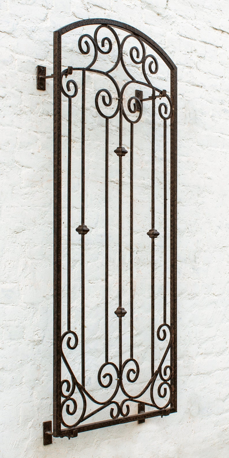 Pu&ograve; includere: Un alto traliccio da parete rettangolare in metallo marrone scuro con una parte superiore ad arco. La struttura &egrave; composta da barre verticali arricchite da piccole sfere decorative e complessi motivi a volute nelle sezioni superiore e inferiore.
