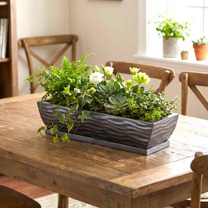 May include: A rectangular, dark gray planter filled with a variety of green plants and white flowers. The planter has a wavy textured design and sits on a wooden table. The arrangement includes ferns, succulents, and ivy.