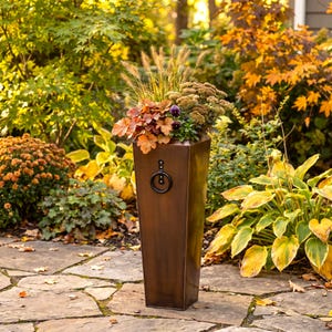 May include: A tall, tapered, bronze-colored metal planter with a dark circular ring handle attached to the front. The planter is filled with a variety of plants, including tall ornamental grasses, small purple flowers, and clusters of brown foliage.