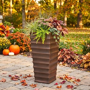 May include: A tall, square, bronze-colored planter with a horizontal ribbed texture. The planter is filled with a variety of plants, including tall green grasses, small pink flower clusters, dark red foliage, and trailing green ivy leaves.