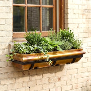 May include: A rectangular copper window box planter with a weathered patina finish, secured by a black metal frame. The planter is filled with a variety of lush green plants, including ivy, hostas, and herbs, positioned beneath a wooden window.