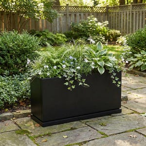 May include: A rectangular black planter box filled with various green plants and white flowers. The planter sits on a stone patio, with a wooden fence and lush greenery in the background. The box is approximately 36 inches long.