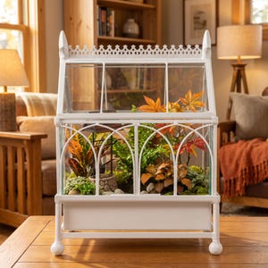 May include: A white metal greenhouse terrarium with a peaked roof and arched window frames. The structure contains various green and orange foliage, including ferns and maple-like leaves, along with a small stone-textured decorative well feature.
