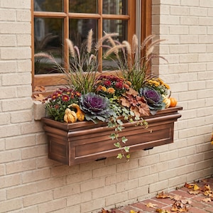 May include: A rectangular bronze-colored window box planter filled with ornamental grasses, deep red and yellow chrysanthemums, purple kale, trailing ivy, and small decorative gourds, mounted against a light-colored brick wall exterior.
