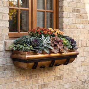 May include: A rectangular copper-toned metal window box planter mounted on a light-colored brick wall. The planter is filled with a variety of plants, including orange and yellow chrysanthemums, purple and green ornamental kale, and ivy.