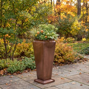 May include: A tall, tapered, copper-colored planter with a square base and rim, filled with a variety of green foliage, tall ornamental grasses, and small orange flowers, standing on a stone patio in an outdoor garden setting.