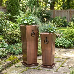 May include: Two tall, copper-colored planters with black ring handles, filled with greenery, stand on a stone patio. The planters have a rectangular base and a tapered, octagonal shape. The background features a lush garden setting.