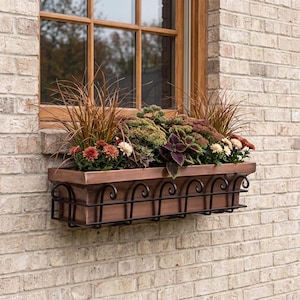 May include: A rectangular copper-colored window box planter featuring a black metal scrollwork cage exterior. The planter is filled with a variety of plants, including tall ornamental grasses, green foliage, and small red, white, and pink flowers.