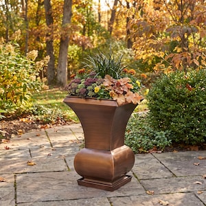 May include: A metallic copper-colored planter with a flared top and a rounded base, filled with a variety of green, orange, and yellow plants including tall grasses, small flowers, and leafy foliage, standing on a stone patio surface.