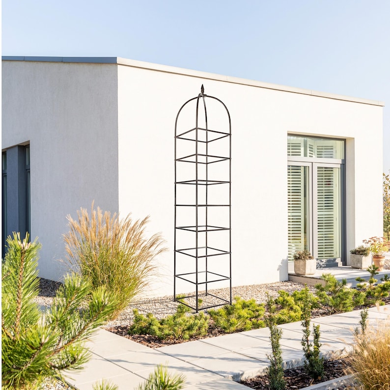 May include: A tall, black metal garden obelisk trellis featuring a square-based frame with horizontal crossbars and a rounded, arched top finished with a decorative finial point. The structure is designed for climbing plants.