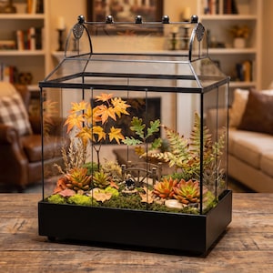 May include: A rectangular glass terrarium with a black metal frame and a peaked roof. Inside, the display features artificial orange maple leaves, green oak leaves, ferns, succulents, moss, small stones, and a miniature arched bridge figurine.