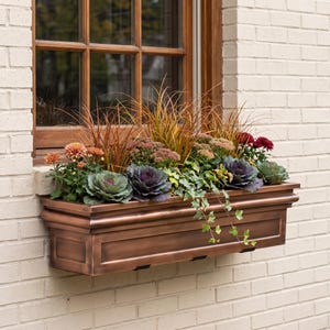 May include: A rectangular copper-toned window box planter mounted beneath a wooden window frame. The planter is filled with a variety of plants, including orange chrysanthemums, purple ornamental kale, tall grasses, and trailing green ivy.